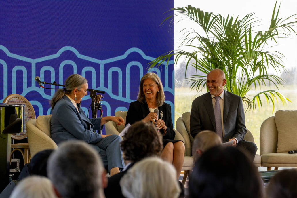 Três convidados sentados em palco, sorrindo e conversando durante um painel com fundo azul e plantas ao redor, diante do público presente.