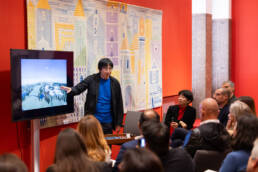 Arquiteto japonês Tezuka apresenta projeto visual em tela, apontando para a imagem enquanto o público observa atentamente no Espaço Santander.