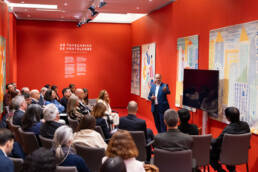 Homem de fato azul faz apresentação para plateia cheia em auditório decorado com tapeçarias da Portalegre e fundo vermelho com o nome da exposição.